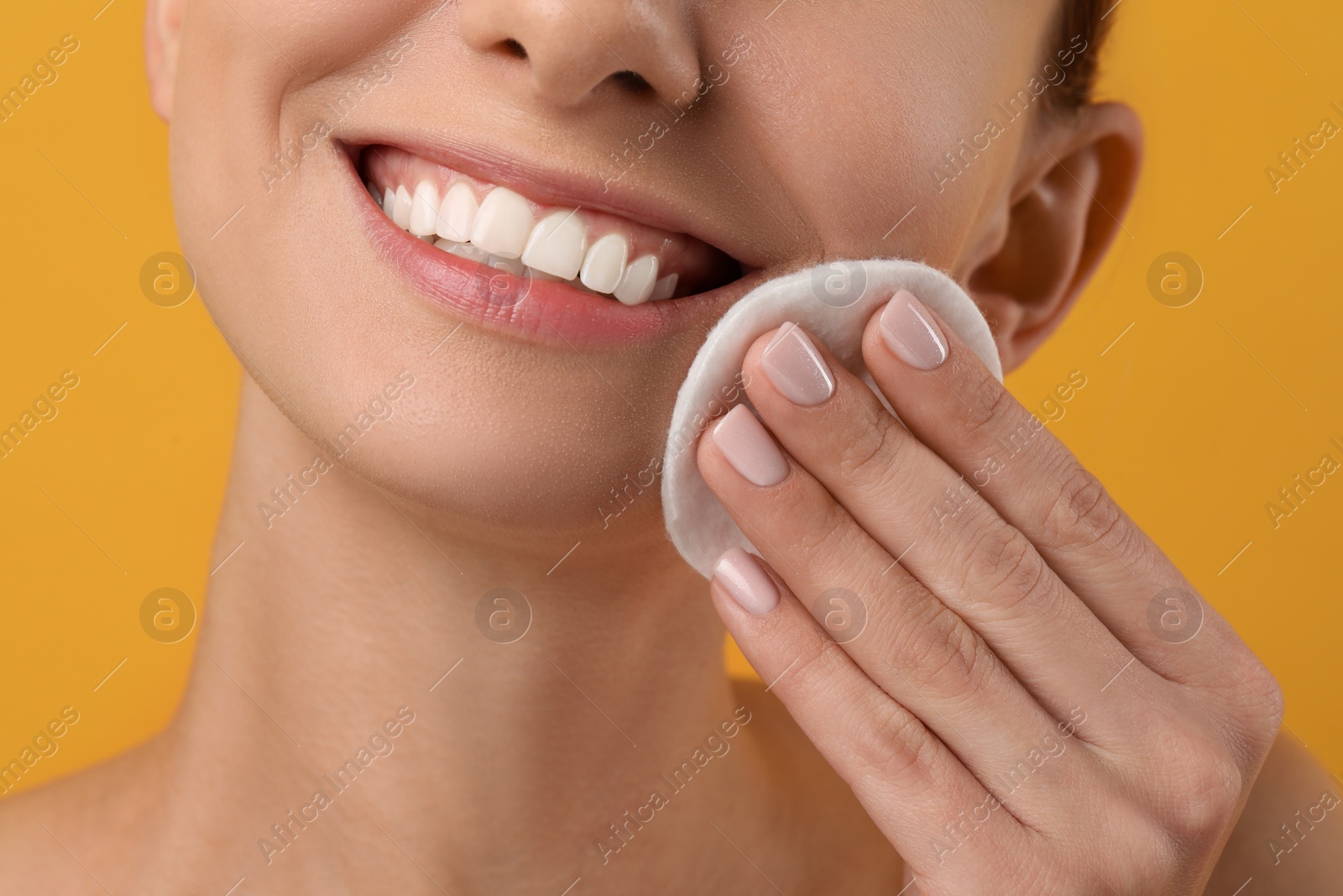 Photo of Smiling woman removing makeup with cotton pad on yellow background, closeup