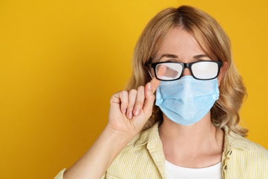 Photo of Woman wiping foggy glasses caused by wearing medical mask on yellow background. Space for text