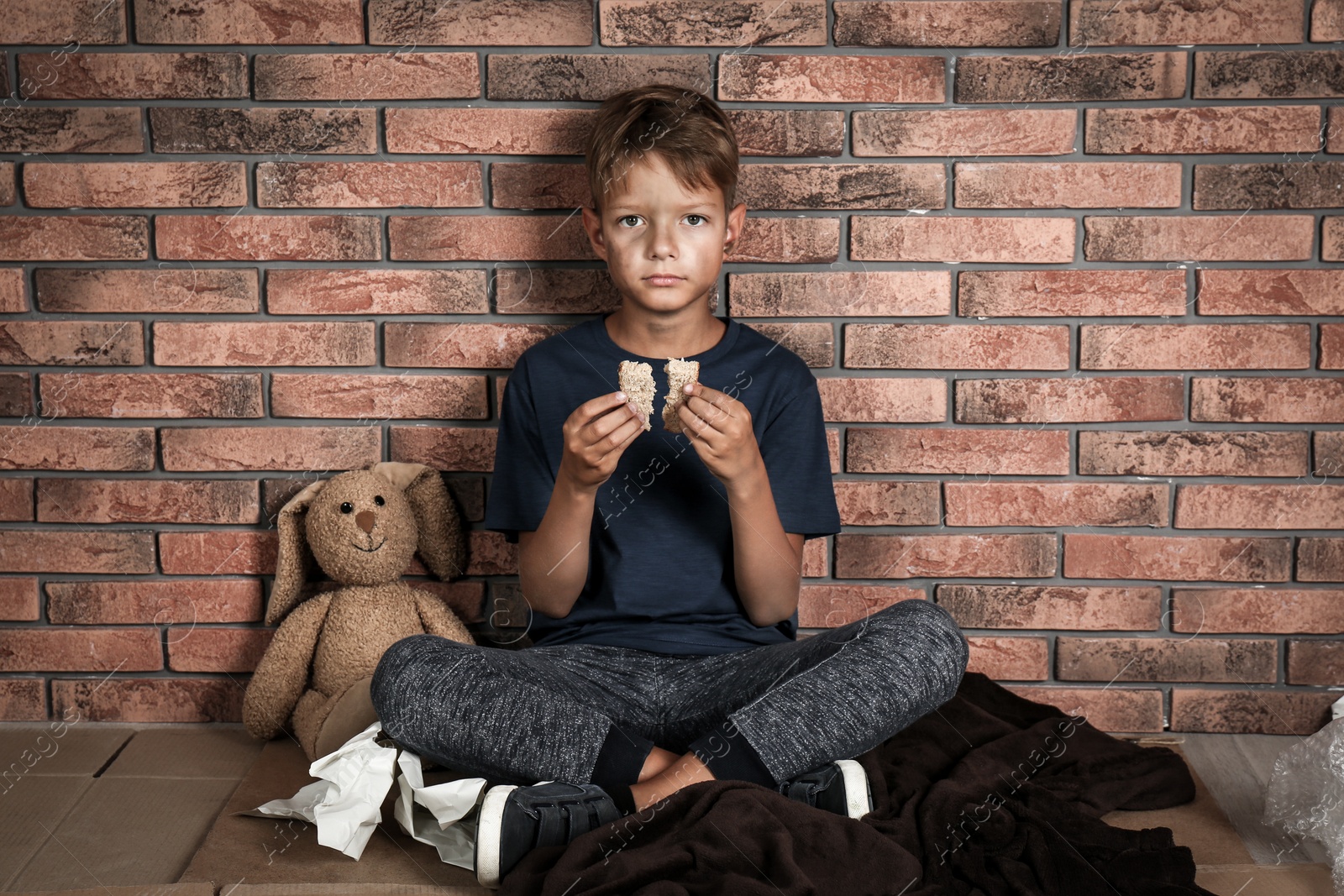 Photo of Poor homeless boy holding bread in hands near brick wall