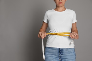 Woman measuring waist with tape on grey background, closeup. Space for text