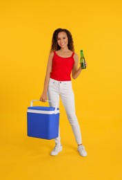 Happy young African American woman with cool box and bottle of beer on yellow background