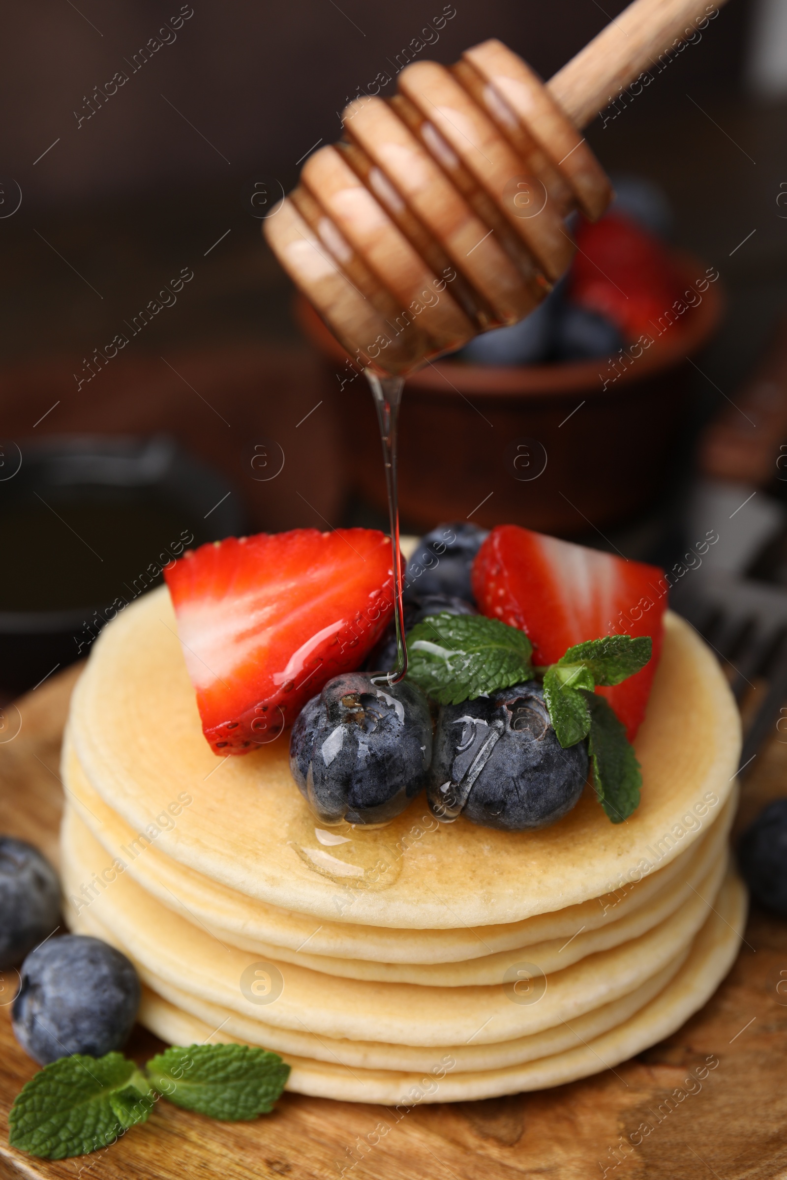 Photo of Pouring honey from dipper onto delicious pancakes with strawberries, blueberries and mint at table, closeup