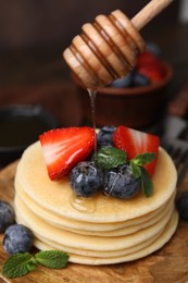 Photo of Pouring honey from dipper onto delicious pancakes with strawberries, blueberries and mint at table, closeup