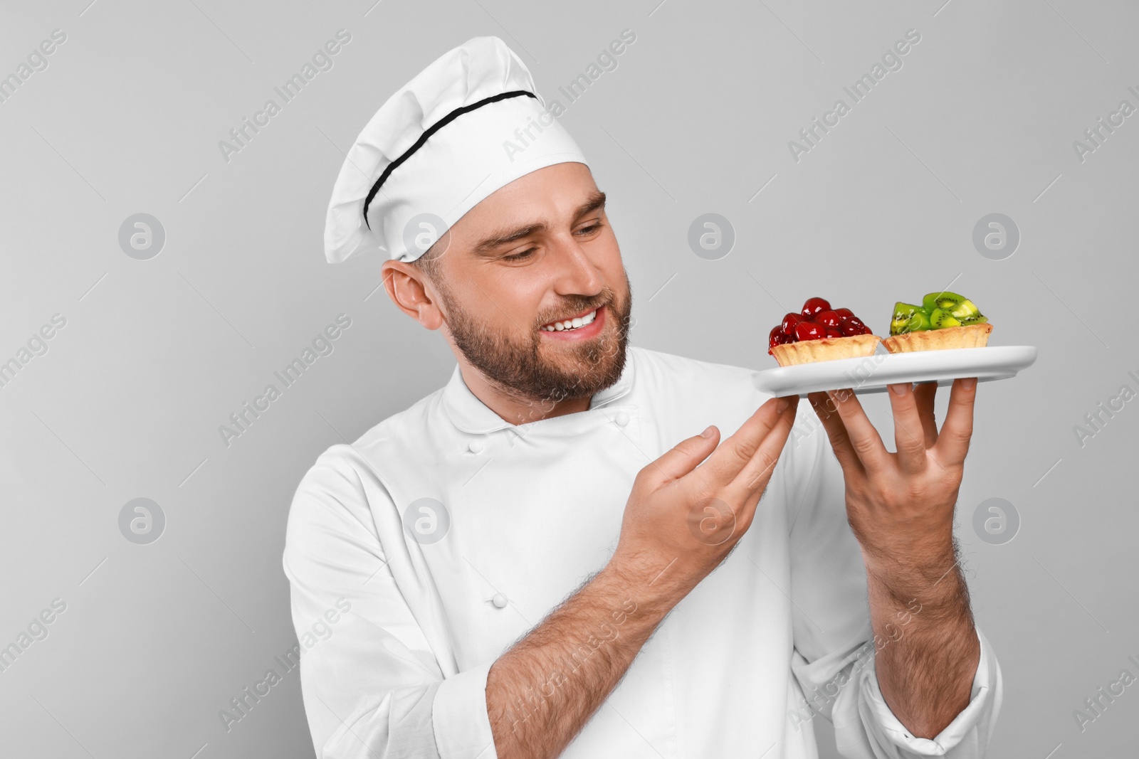 Photo of Happy professional confectioner in uniform holding plate with delicious tartlets on light grey background