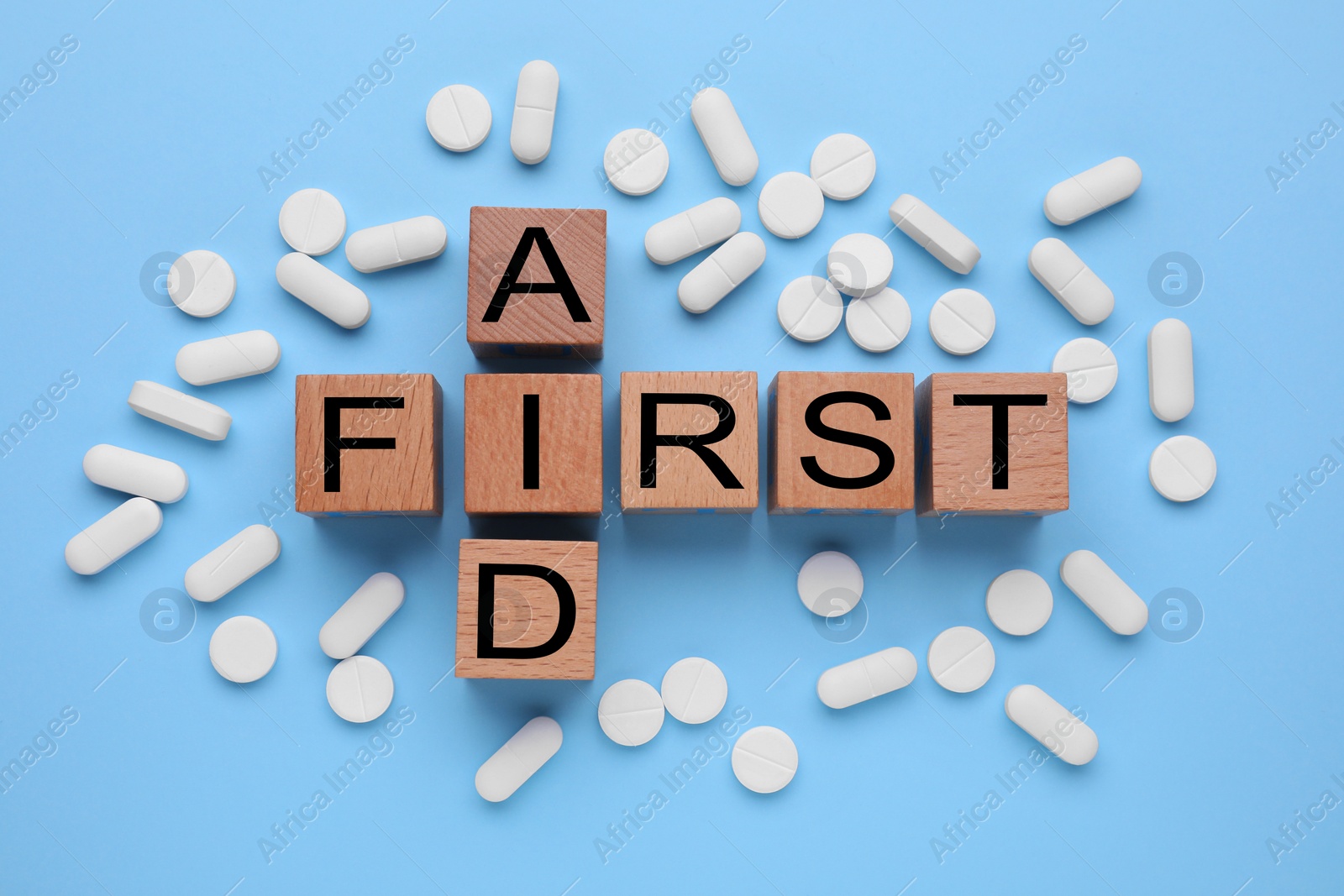 Photo of Words First Aid made of wooden cubes and pills on light blue background, flat lay