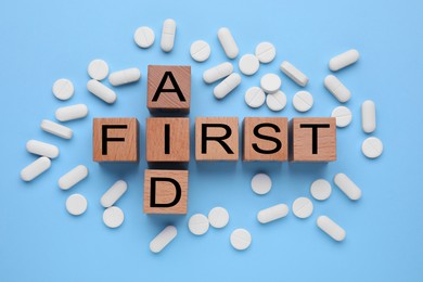 Photo of Words First Aid made of wooden cubes and pills on light blue background, flat lay