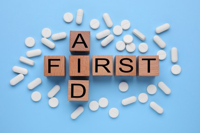 Words First Aid made of wooden cubes and pills on light blue background, flat lay