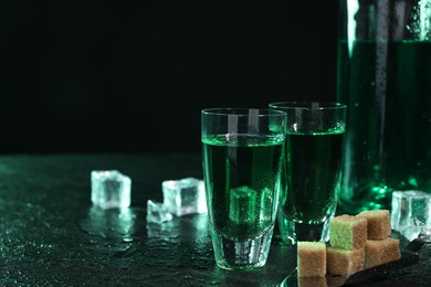 Absinthe in shot glasses, spoon, brown sugar and ice cubes on gray table against dark background, space for text. Alcoholic drink