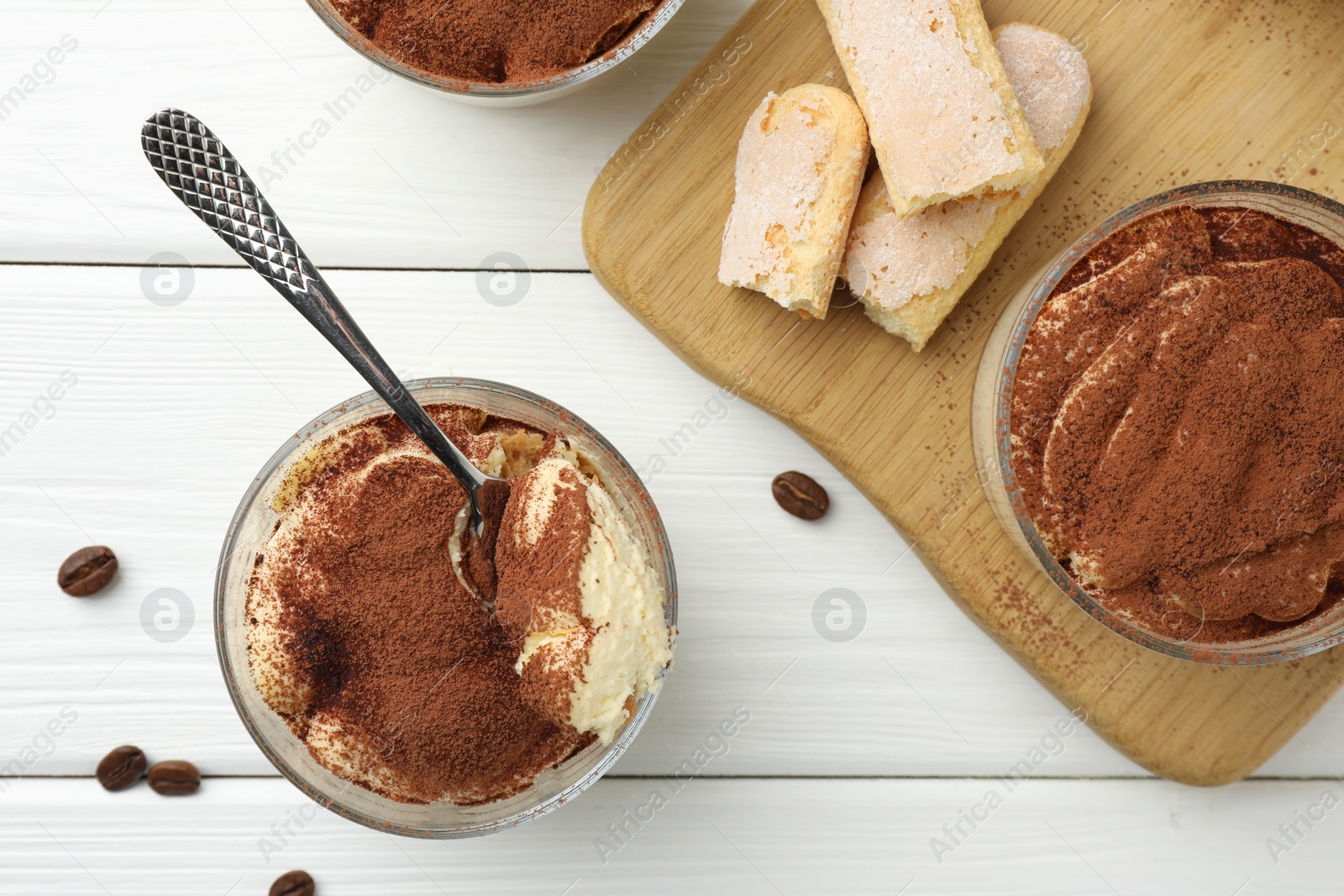 Photo of Delicious tiramisu in glasses, spoon, coffee beans and cookies on white wooden table, top view