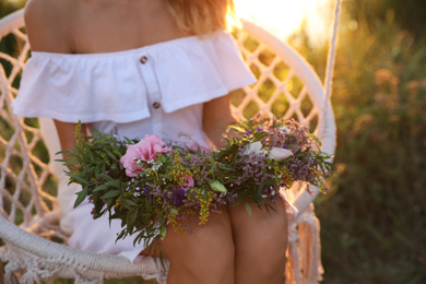 Photo of Young woman holding wreath made of beautiful flowers on swing chair outdoors at sunset, closeup
