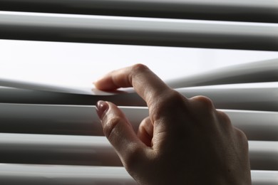 Photo of Woman separating slats of white blinds indoors, closeup