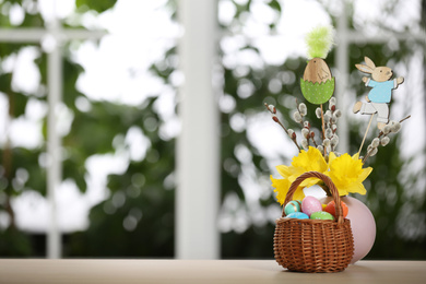 Photo of Festive composition with Easter eggs in wicker basket on table against blurred window, space for text