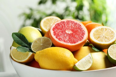 Photo of Different fresh citrus fruits and leaves in bowl against blurred background, closeup