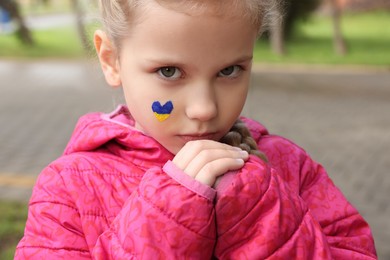 Photo of Little girl with drawing of Ukrainian flag on face in heart shape outdoors, closeup