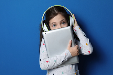 Photo of Cute little girl with headphones and tablet listening to audiobook on blue background