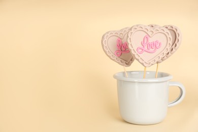 Photo of Chocolate heart shaped lollipops with word Love in cup on beige background. Space for text