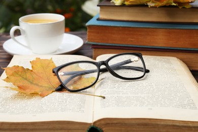 Photo of Yellow maple leaf, glasses, book and cup of tea on wooden table, closeup. Autumn atmosphere