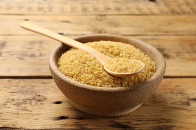 Photo of Bowl and spoon with raw bulgur on wooden table, closeup