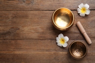 Photo of Golden singing bowls, mallet and flowers on wooden table, flat lay. Space for text