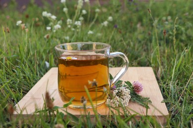 Photo of Cup of aromatic herbal tea and different wildflowers on wooden board in meadow