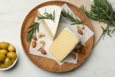 Photo of Plate with pieces of tasty camembert cheese, nuts, olives and rosemary on white marble table, flat lay