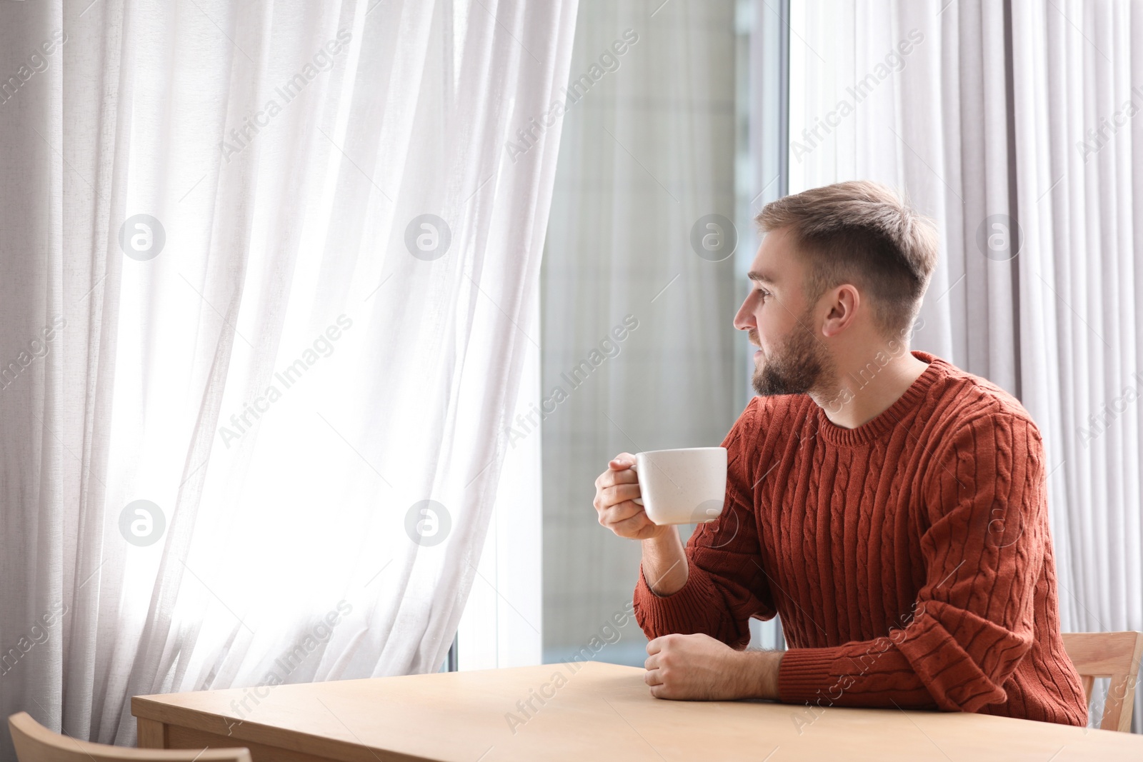 Photo of Young man with cup of hot drink near window at cafe