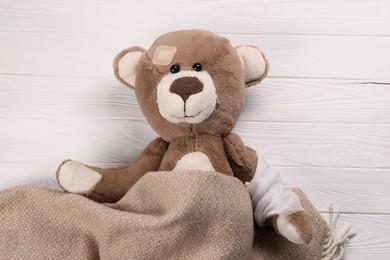 Photo of Toy bear with bandage and medical plaster under blanket on wooden background, top view